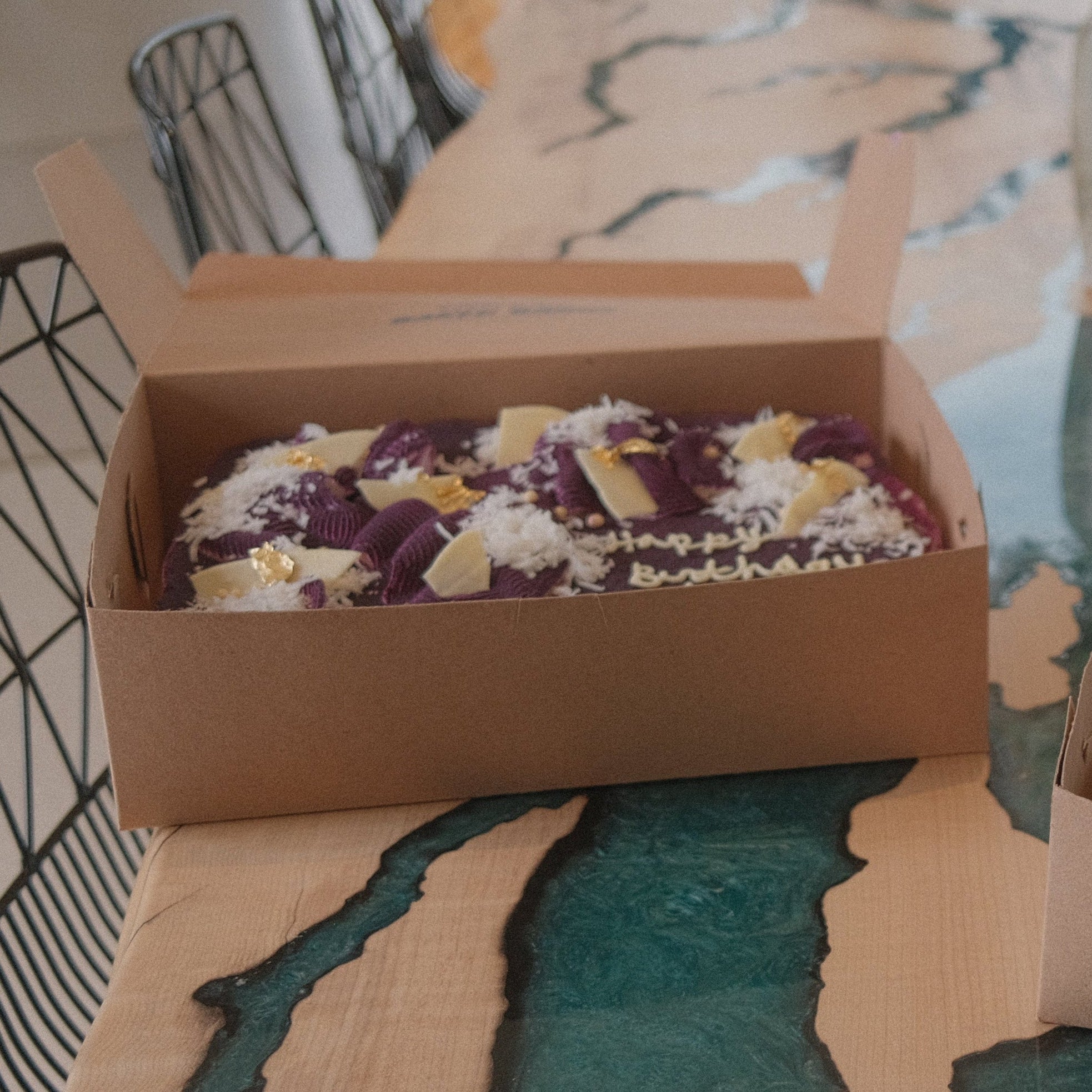 A whole ube genoise cake in a cardboard box on a kitchen island with blue resin designs. 